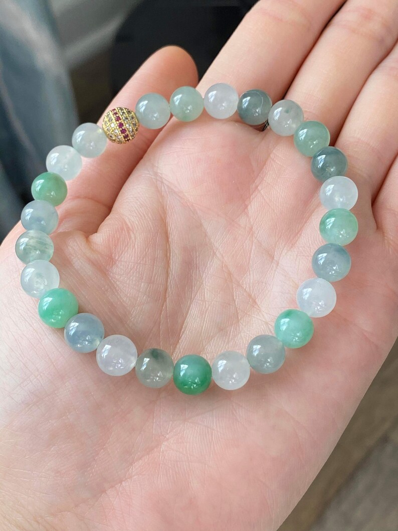 Pode incluir: Um bracelete de jade&iacute;ta com uma conta de ouro e pedra vermelha. O bracelete &eacute; feito de contas redondas em tons de verde e branco.