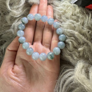 May include: A light blue jade bracelet with green accents. The bracelet is made of round beads and is strung on a clear elastic cord.