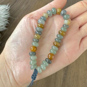 Könnte beinhalten: Ein Armband aus abgerundeten, mehrfarbigen Perlen in Grün-, Braun- und Grautönen. Die Perlen sind auf einem grauen Band mit einem geknoteten Verschluss aufgefädelt. Das Armband wird in einer Hand gehalten.