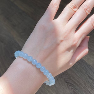 K&ouml;nnte beinhalten: Ein hellblaues Perlenarmband, das an einem Handgelenk getragen wird. Das Armband besteht aus runden, glatten Perlen.