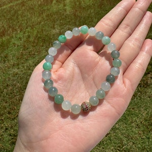 Pode incluir: Um bracelete de contas de jade verde e branco com uma conta de destaque de pedra preciosa dourada e vermelha.