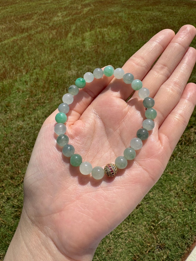 Pode incluir: Um bracelete de contas de jade verde e branco com uma conta de destaque de pedra preciosa dourada e vermelha.