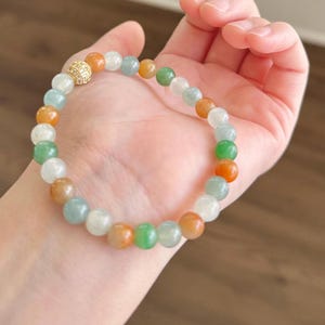 May include: A jade bracelet with a gold bead accent. The bracelet is made of round beads in shades of green, white, and orange.