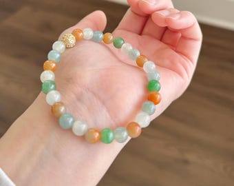 Pode incluir: Um bracelete de jade com um detalhe de contas douradas. O bracelete é feito de contas redondas em tons de verde, branco e laranja.