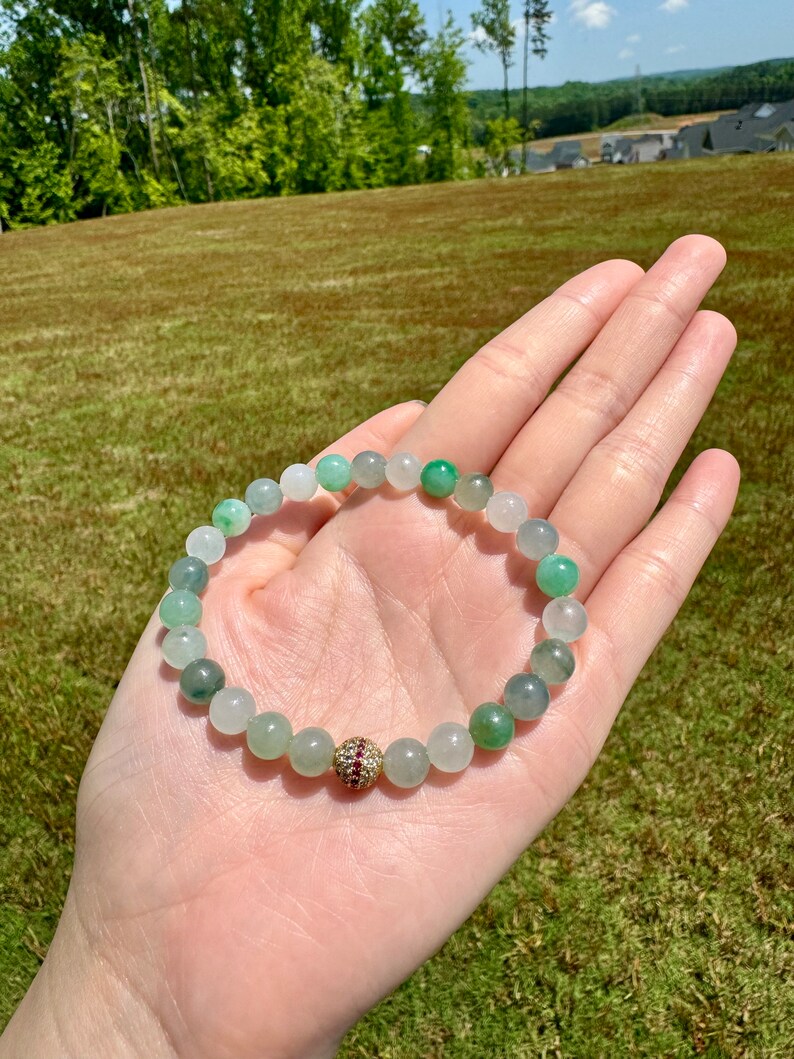 Pode incluir: Um bracelete de contas de jade verde e branco com uma conta de destaque de pedra preciosa dourada e vermelha.