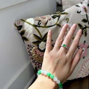 May include: A green and white jade bead bracelet with a silver ring with a green gemstone on the finger.
