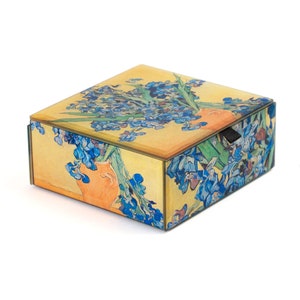 May include: A square-shaped decorative box with a hinged lid. The box is made of glass and features a colorful image of a vase of blue irises. The box has a black pull tab on the side.