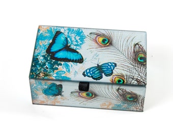 Puede incluir: Una caja de vidrio rectangular para baratijas con tapa con bisagras. La caja está decorada con un diseño colorido de mariposas azules y plumas de pavo real sobre un fondo azul claro.