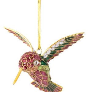 May include: A gold hummingbird ornament with pink, green, and white enamel detailing. The hummingbird is in flight with its wings spread out.