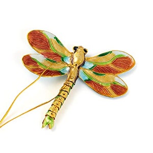 May include: A decorative dragonfly ornament with a gold-colored body and wings. The wings feature a detailed design with shades of brown, green, and gold. A thin gold string is attached for hanging. The ornament is likely made of metal.