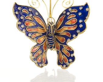 May include: A decorative butterfly ornament with blue, orange, and gold accents. The butterfly has a detailed design with small, clear embellishments along the edges of the wings. It hangs from a gold-colored string.