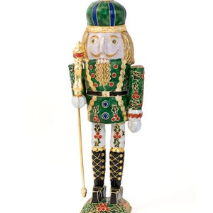 Puede incluir: Una figurita de cascanueces pintada de verde y dorado con un patrón de acebo rojo y blanco. El cascanueces lleva una corona verde y azul y sostiene un bastón dorado.