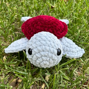 May include: A crocheted plush toy turtle with a gray body and red shell. The turtle is sitting on a bed of green grass.