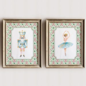 Puede incluir: Dos impresiones en acuarela enmarcadas. Una presenta un soldado cascanueces azul y dorado. La otra representa a una bailarina con tutú azul y corona dorada. Ambas impresiones tienen un borde floral y marcos dorados.