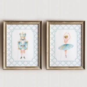 May include: Two framed watercolor prints. One features a blue and gold nutcracker soldier. The other depicts a ballerina in a pink top and blue tutu. Both prints have a light blue patterned border and gold frames.