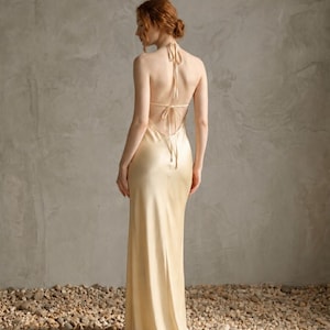 May include: A floor-length, champagne-colored satin dress featuring a backless design with thin, adjustable tie-up straps at the neck and mid-back. The garment has a smooth, lustrous finish and a fluid, straight-cut silhouette that falls to the floor.