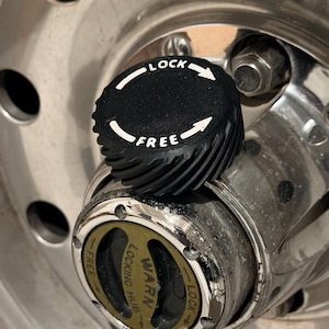 May include: A black locking hub cap with white text that reads "LOCK" and "FREE" with directional arrows. The hub cap is on a silver wheel. The hub cap has a ridged edge.
