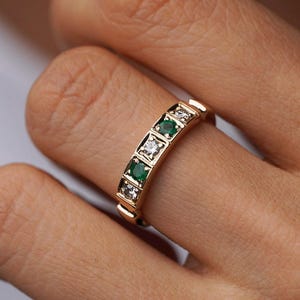 May include: A gold ring with a series of square-cut emeralds and diamonds. The ring is set in a gold band. The gemstones are arranged in a repeating pattern, alternating between green emeralds and clear diamonds. The ring is worn on a finger.