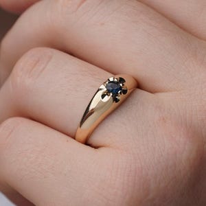 May include: A gold ring with a dark blue gemstone set in a starburst design. The ring is smooth and polished, with a classic, elegant style. The gemstone is small and round, adding a touch of color to the gold band.