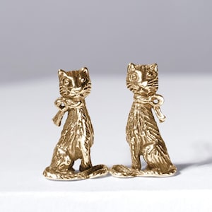 Puede incluir: Dos figuras de gato doradas. Cada gato está sentado y lleva un lazo alrededor del cuello. Los gatos tienen detalles, incluyendo bigotes y pelaje texturizado. Las figuras se muestran sobre un fondo neutro.