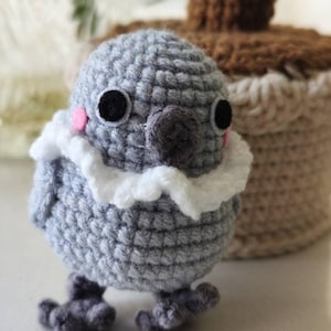 Pode incluir: Uma pequena figura de pássaro em crochê, redonda e em tons de cinza. Apresenta um colarinho branco com babados, dois olhos circulares pretos e pequenos círculos de feltro rosa nas bochechas. O pássaro tem bico e pés de crochê cinza-escuro.