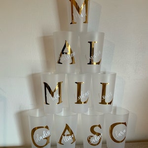May include: Eight frosted plastic cups with gold letters and white names on them. The letters spell out the word "MALCAS", with the names "Mila", "Lukas", "Louis", "Gladys", "Alice", "Soline", and "Lara".