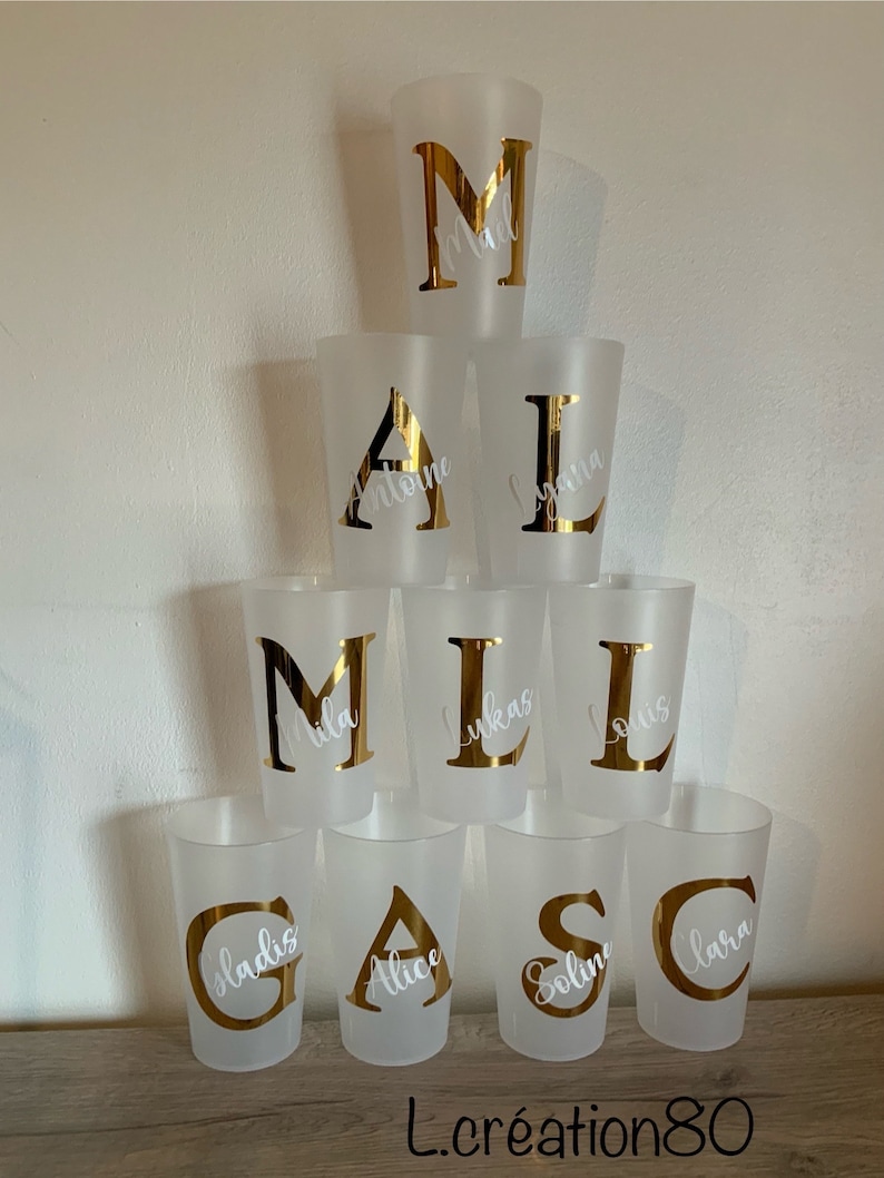 May include: Eight frosted plastic cups with gold letters and white names on them. The letters spell out the word "MALCAS", with the names "Mila", "Lukas", "Louis", "Gladys", "Alice", "Soline", and "Lara".