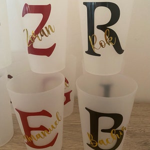 May include: Four clear plastic cups with red and black letters and gold lettering. The letters are Z, R, E, and B. The names on the cups are "Zoran", "Rok", "Emmanuel", and "Bachir".