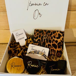 Könnte beinhalten: Eine weiße Geschenkbox gefüllt mit Accessoires, darunter eine Geldbörse mit Leopardenmuster, ein Haargummi und eine Sonnenbrille. Die Box trägt den goldenen Schriftzug "Témoin en Or" und enthält einen Kompaktspiegel und herzförmige Sonnenbrillen.