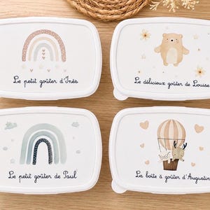 Peut inclure: Quatre boîtes à goûter rectangulaires blanches aux coins arrondis. En haut à gauche, un arc-en-ciel et "Le petit goûter d'Inès". En haut à droite, un ours et "Le délicieux goûter de Louise". En bas à gauche, un arc-en-ciel et "Le petit goûter de Paul". En bas à droite, une montgolfière et "La boîte à goûter d'Augustin".