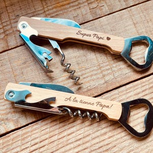 Könnte beinhalten: Zwei Weinöffner aus Holz mit Edelstahlkomponenten. Auf einem Öffner steht der Text "Super Papi". Auf dem anderen Öffner steht der Text "A la tienne Papa!". Beide Werkzeuge sind Flaschenöffner.
