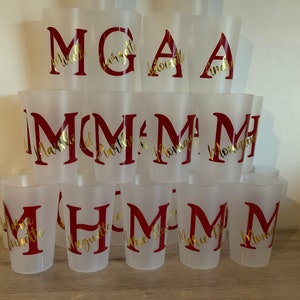 May include: A stack of clear plastic cups with gold lettering on a wooden surface. The cups are decorated with the letters M, G, and A, and the names of various people.