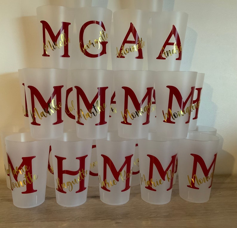May include: A stack of clear plastic cups with gold lettering on a wooden surface. The cups are decorated with the letters M, G, and A, and the names of various people.