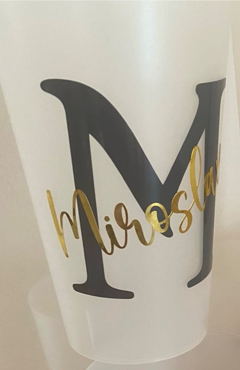 May include: A white plastic cup with a black letter "M" and the name "Miroslav" written in gold cursive lettering.