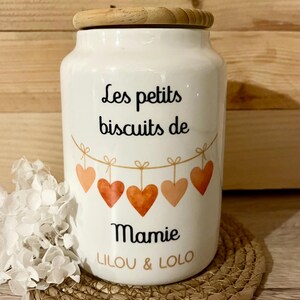 Peut inclure: Pot en céramique blanc avec un couvercle en bois, portant l'inscription "Les petits biscuits de Mamie" et une guirlande de cœurs. Le pot est posé sur un tapis tressé.