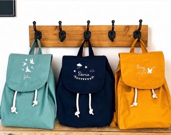 Könnte beinhalten: Drei Rucksäcke in Salbeigrün, Marineblau und Senfgelb, jeweils mit einer Klappe, die mit weißen Stickereien und Namen versehen ist. Der salbeigrüne Rucksack zeigt Vögel, der marineblaue Wolken und den Namen Léonie, der gelbe einen Schmetterling.