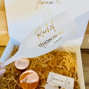 Peut inclure: Une flûte à champagne givrée avec le texte doré Rachel, TÉMOIN EN OR et 13.06.2026. Dans une boîte cadeau blanche avec de la paille de bois se trouvent un miroir de poche rond en or rose avec une rose et un bracelet blanc avec un cœur noir.
