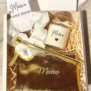 Può includere: Una confezione regalo contenente una penna dorata, una pochette dorata, una candela e uno scrunchie bianco. La penna ha una parte superiore a forma di cuore. La candela e la pochette sono iscritte con il nome "Maëva". La scatola include anche una carta con "Maëva SUPER TEMOIN".