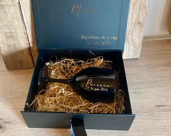May include: A black gift box with a black ribbon and a black wine glass with gold lettering that says "Parrain 16 juin 2024" inside. The box lid has gold lettering that says "Baptême de Leila 16 juin 2024".