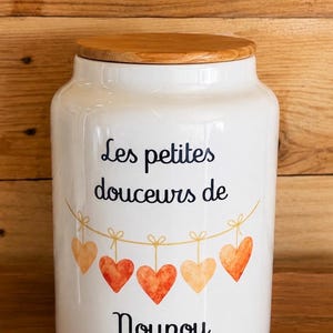 Peut inclure: Un bocal de rangement en céramique blanche avec un couvercle en bois. L'avant présente le texte noir "Les petites douceurs de Nounou" au-dessus d'une guirlande de cinq cœurs orange et rouges, avec le nom "MAXENCE" imprimé en doré en dessous.