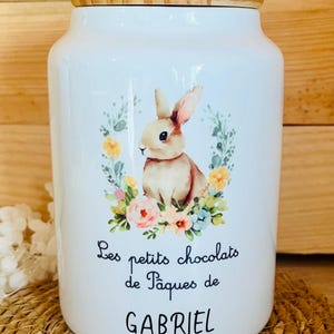 Peut inclure: Pot en céramique blanche avec un couvercle en bois, orné d'une illustration d'un lapin entouré de fleurs. Le pot porte l'inscription "Les petits chocolats de Pâques de GABRIEL".