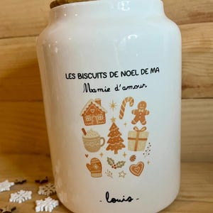 Peut inclure: Un pot en céramique blanc avec un couvercle en bois, portant l'inscription "LES BISCUITS DE NOEL DE MA Mamie d'amour" et "louis" en noir, orné d'un motif graphique coloré de maison en pain d'épices, sucre d'orge, sapin et cadeau.