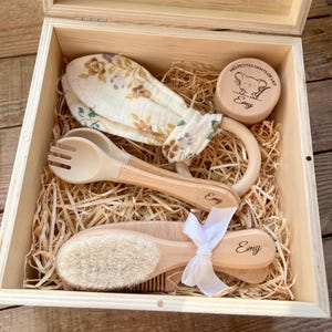 Peut inclure: Un coffret cadeau en bois contenant un ensemble pour bébé avec un anneau de dentition en mousseline fleurie, une fourchette et une cuillère en bois, une boîte ronde gravée d'une souris et du nom Emy, et une brosse à cheveux en bois.