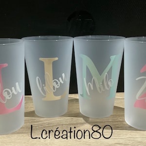 May include: Four frosted plastic cups with personalized names, "Lou", "Lilou", "Milo", and "Zoe", in different colors. The cups are on a wooden surface.