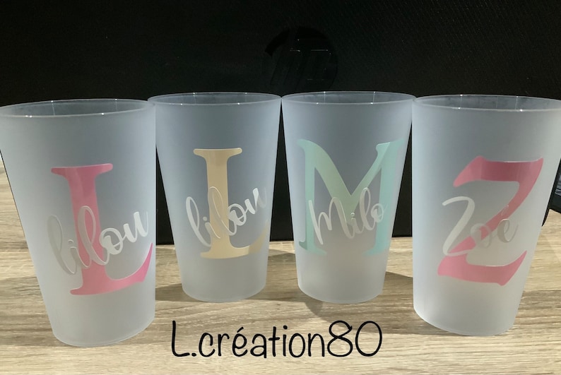 May include: Four frosted plastic cups with personalized names, "Lou", "Lilou", "Milo", and "Zoe", in different colors. The cups are on a wooden surface.