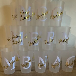 May include: A collection of frosted plastic cups with gold lettering. The cups are stacked in a pyramid shape and feature names such as Marie, Therese, and Christian.