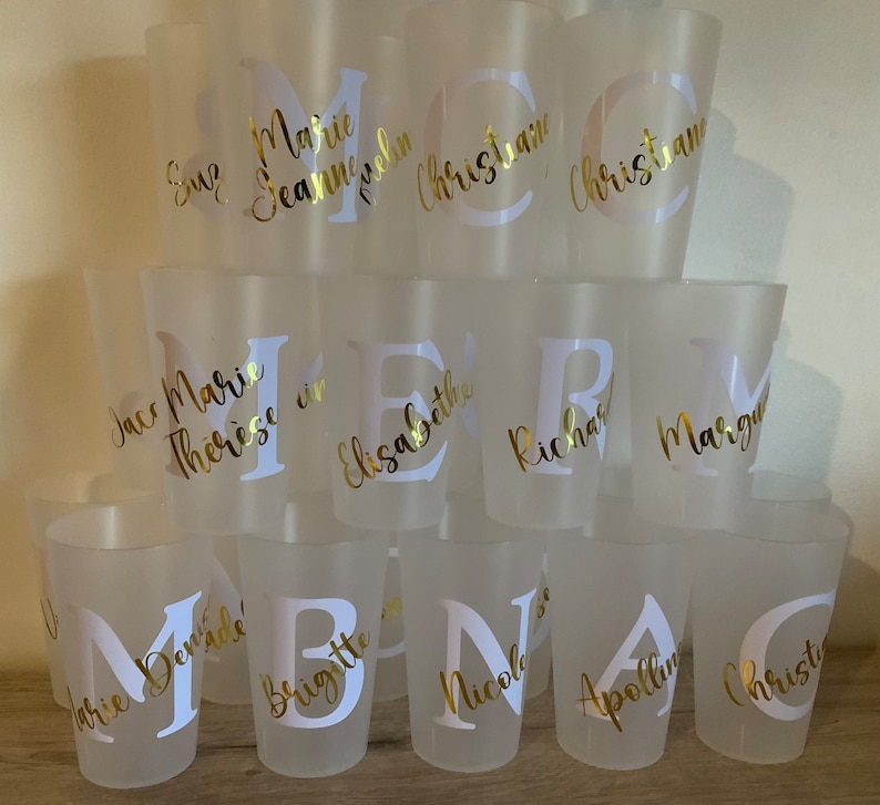 May include: A collection of frosted plastic cups with gold lettering. The cups are stacked in a pyramid shape and feature names such as Marie, Therese, and Christian.