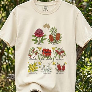May include: A cream-coloured t-shirt displaying botanical illustrations of Australian flowers, such as Waratah and Banksia. The design includes the text "COMFORT". The shirt is shown outdoors, set against a backdrop of green foliage.