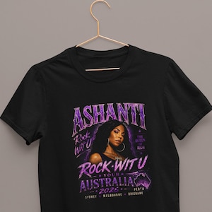 May include: A black short-sleeved t-shirt featuring a purple graphic print of Ashanti. The text reads ASHANTI, ROCK WIT U, THE QUEEN OF R&B, TOUR AUSTRALIA 2025, SYDNEY, MELBOURNE, BRISBANE, and PERTH, along with a map of Australia and star accents.