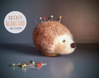 May include: A needle felted brown and cream hedgehog pin cushion with a black nose and eye, featuring several colorful pins inserted into its back. The text PRICKLY HEDGEHOG PIN CUSHION is visible in a white circle. Loose sewing pins lie nearby.